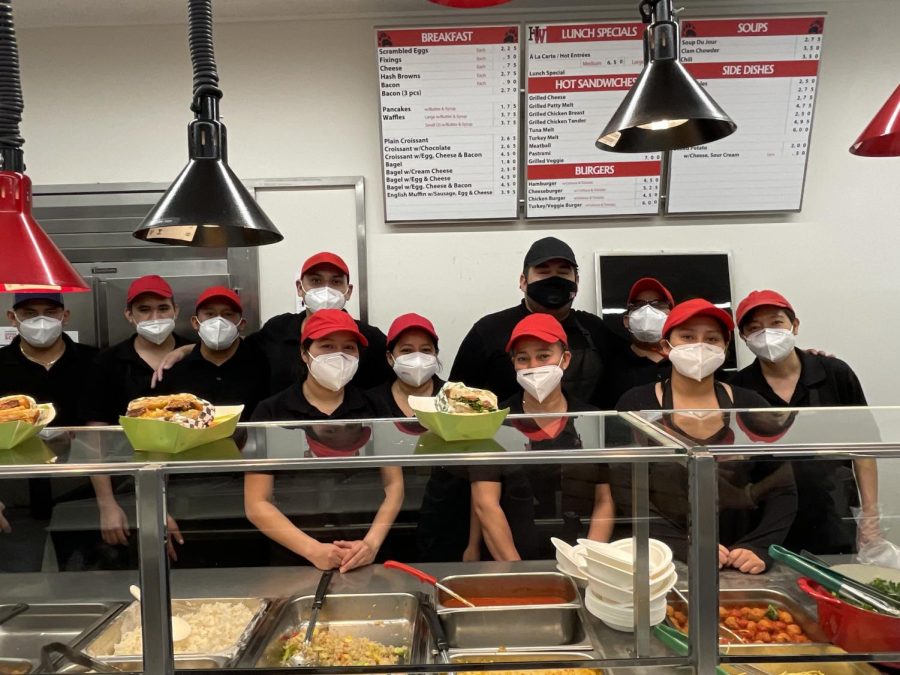 The cafeteria staff stands smiling for a photo in front of the food.
